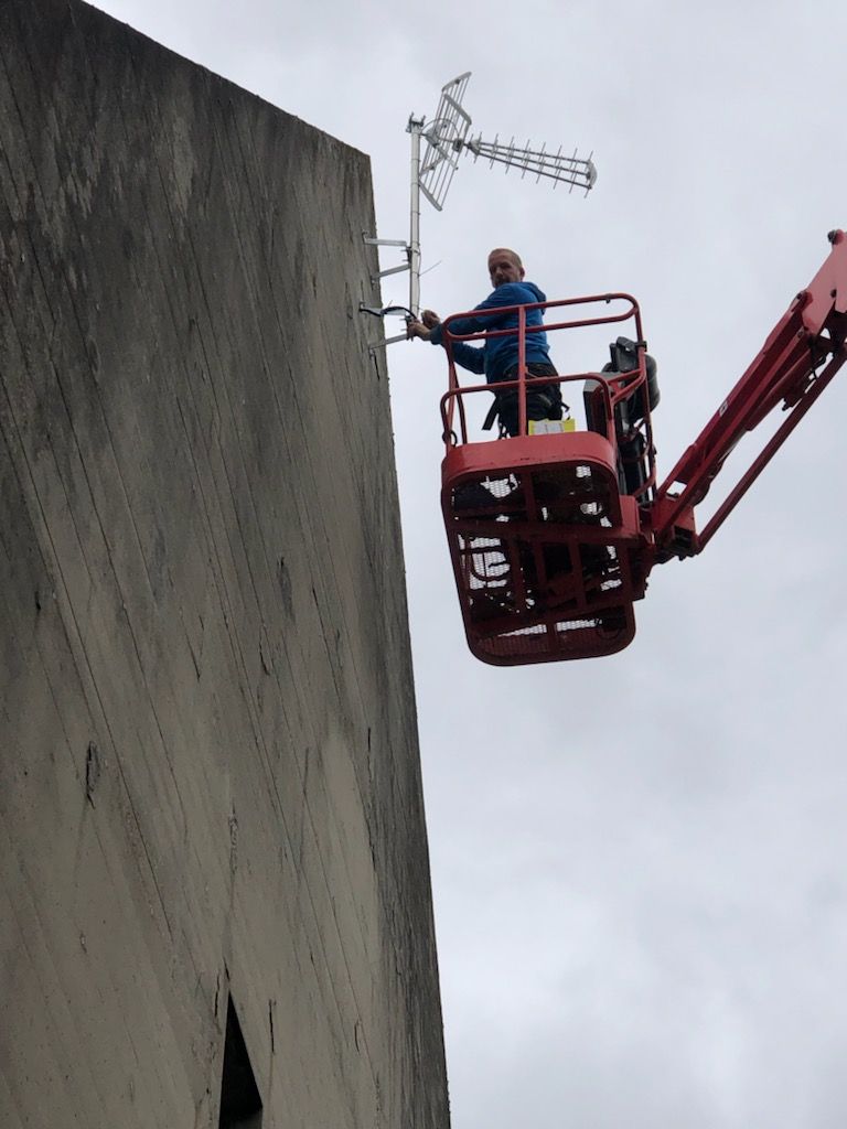 Un ouvrier en veste bleue, sur une nacelle rouge, taille une antenne près d'un haut mur en béton.