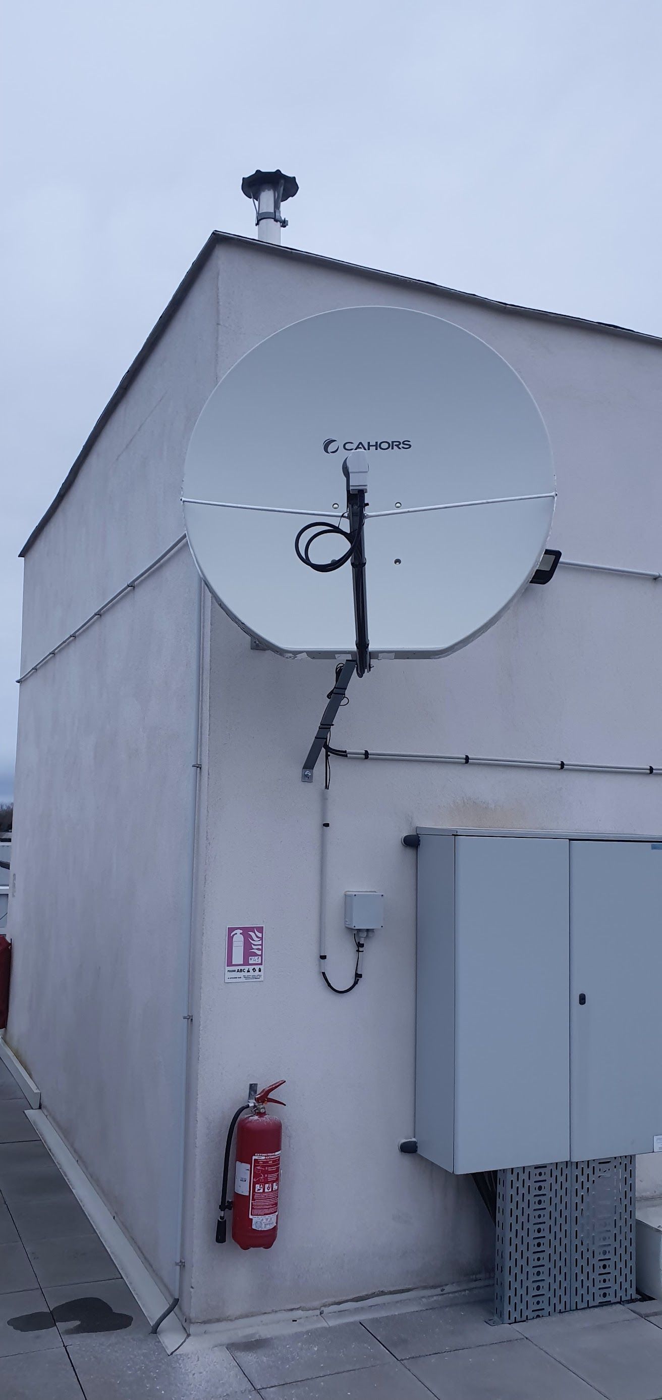 Une grande antenne parabolique blanche est fixée sur le mur gris d'un bâtiment, avec un extincteur rouge en dessous.