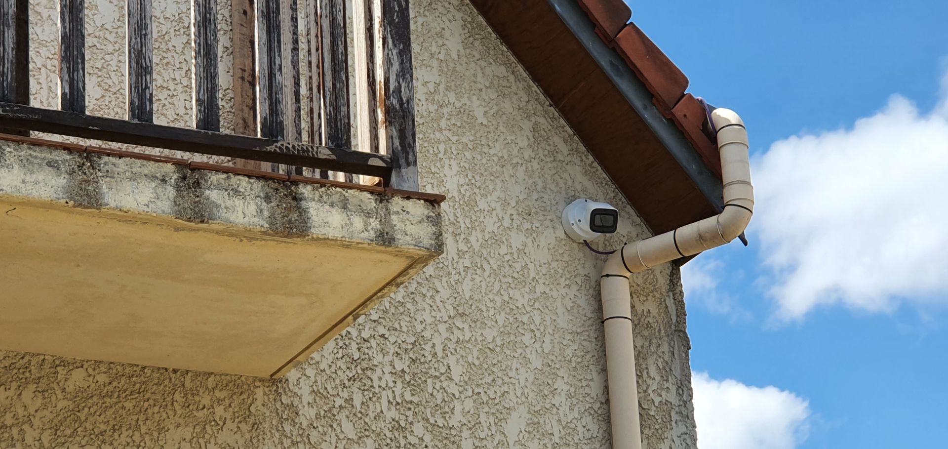 Coin d'un immeuble en stuc avec balcon, descente de gouttière, caméra de sécurité et ciel bleu