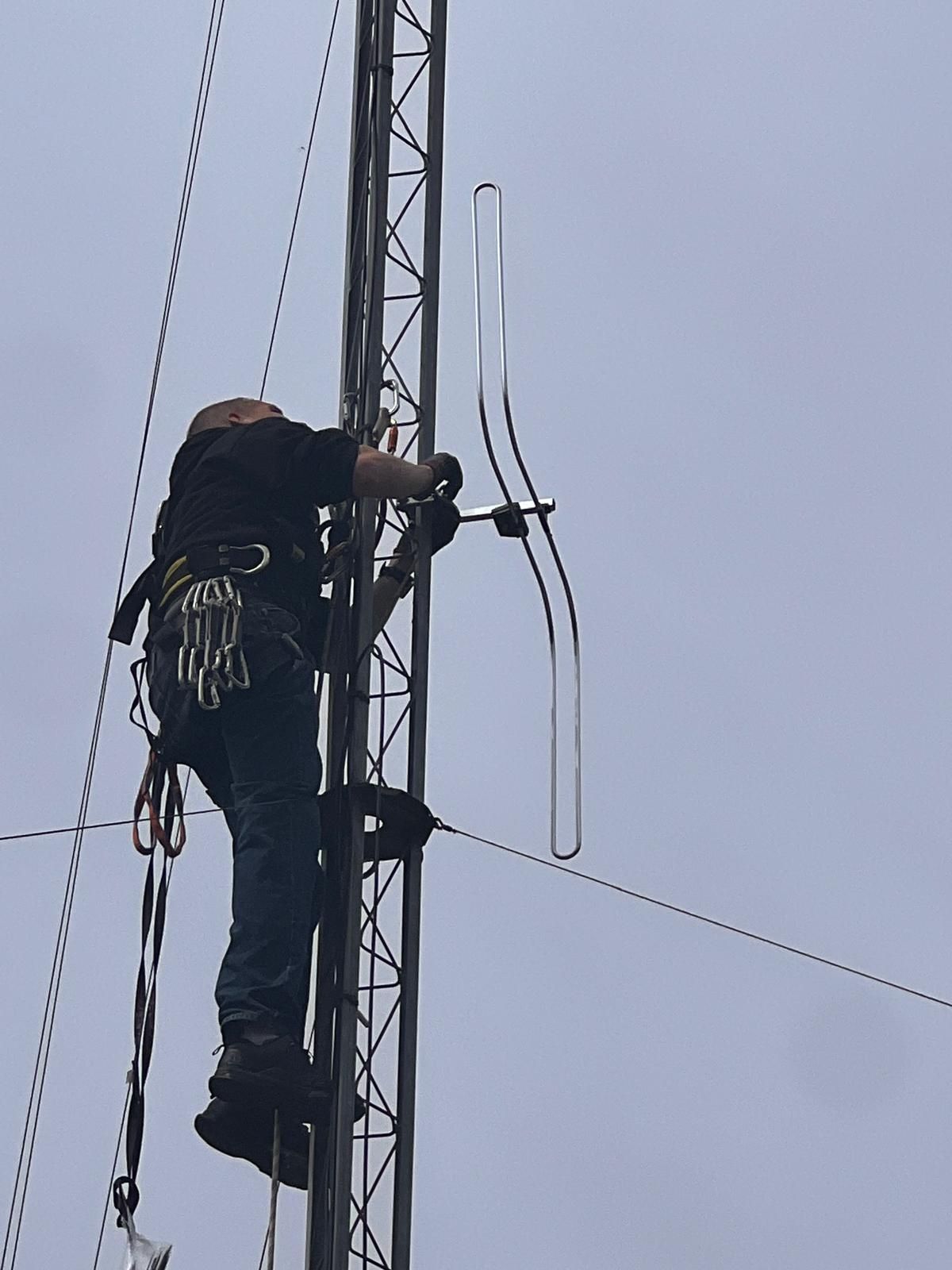 Un ouvrier escalade une tour de télécommunications, ajustant les câbles d'antenne sous un ciel nuageux.