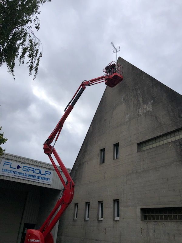 Une nacelle élévatrice rouge soulève un ouvrier pour inspecter le sommet d'un bâtiment industriel gris.