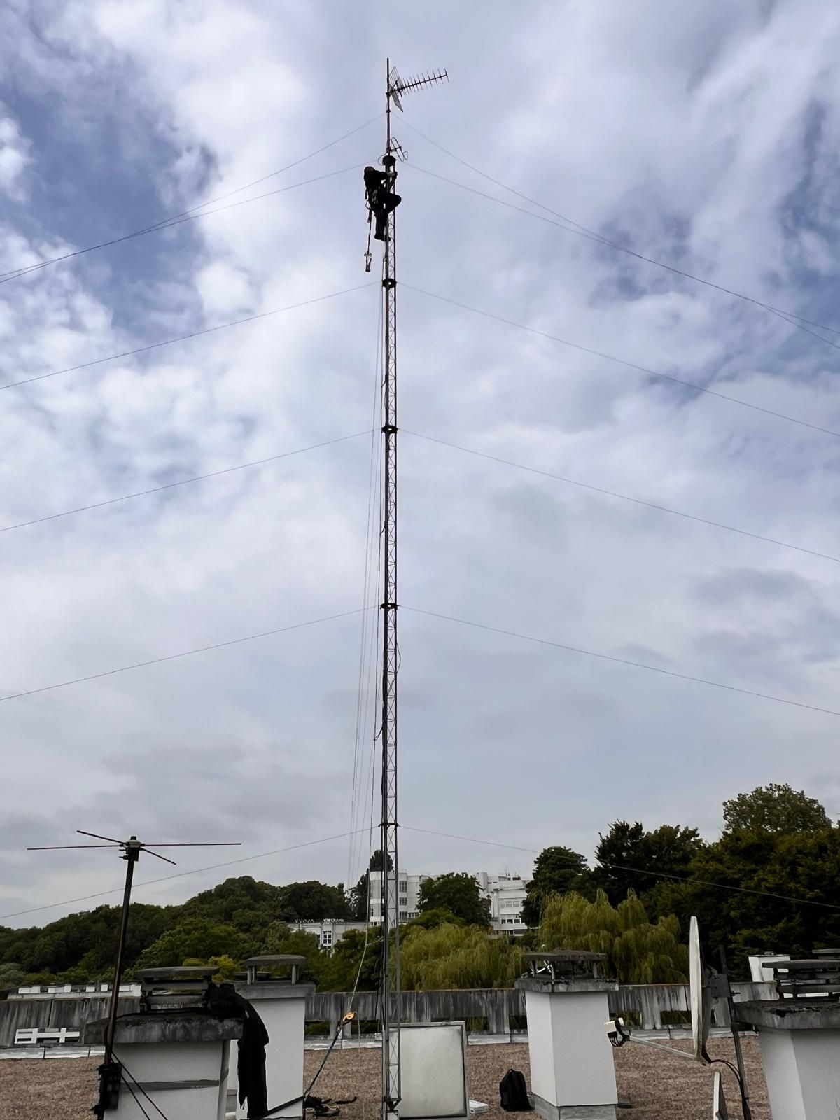 Haute tour de télécommunications avec ses antennes se détachant sur un ciel nuageux, au-dessus de petits bâtiments utilitaires et d'arbres.