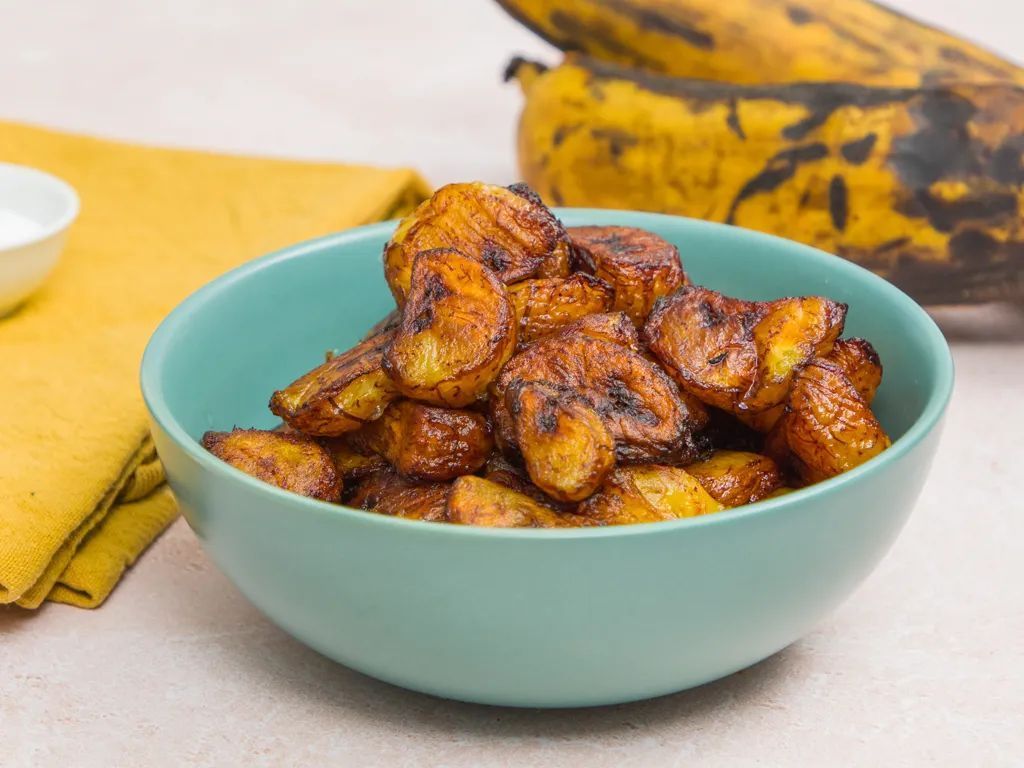 Bananes plantains frites dans un bol turquoise, avec des bananes plantains entières en arrière-plan. Serviette jaune et petit bol blanc de sel.