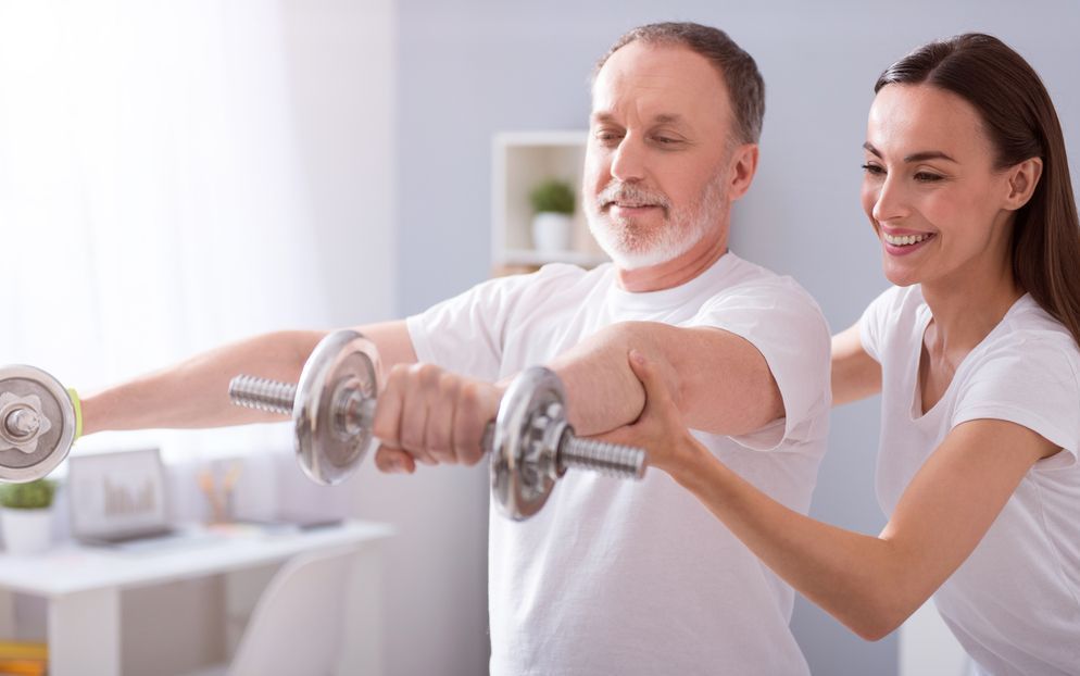 älterer Mann hebt Gewichte und wird von einer Physiotherapeutin unterstützt