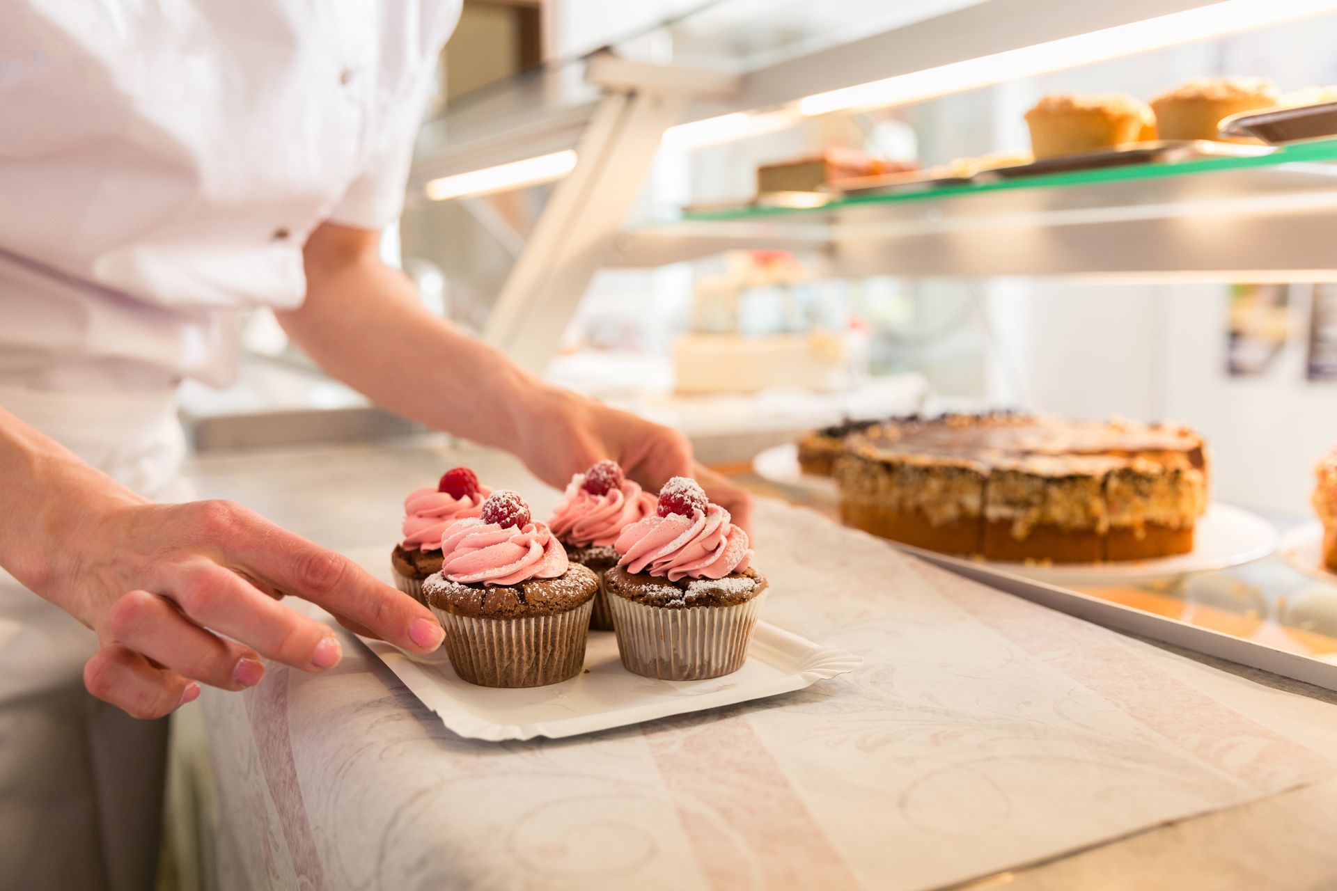 Eine Person in weisser Uniform stellt drei Cupcakes mit rosa Zuckerguss auf eine Theke in einer Bäckerei.