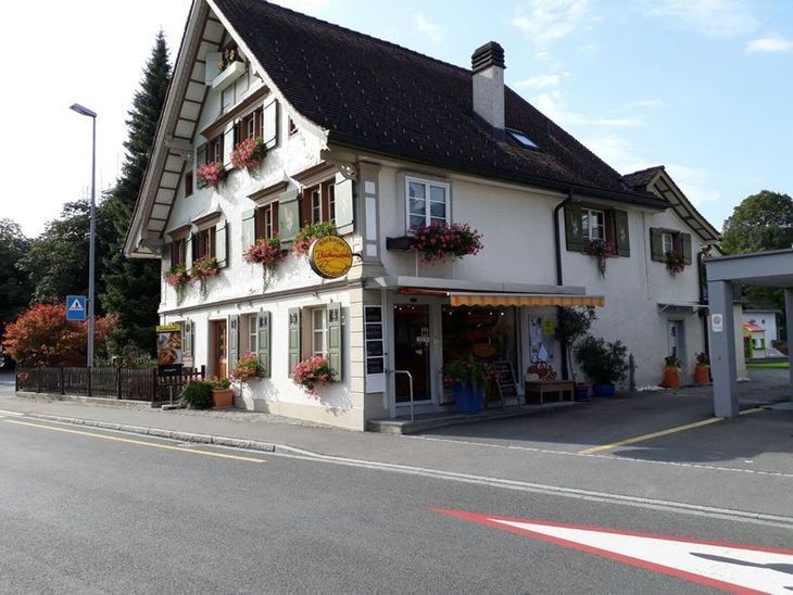 Eckgebäude mit weissen Wänden, braunen Fensterläden, Blumenarrangements und einem Schild.