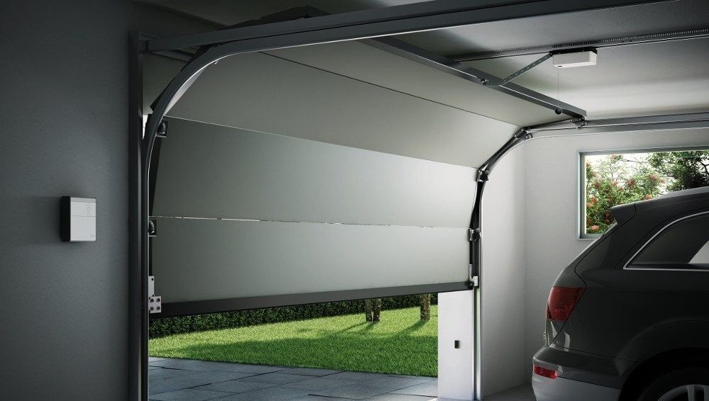 Una moderna puerta de garaje, entreabierta, deja ver un césped, con un coche oscuro aparcado en el interior.