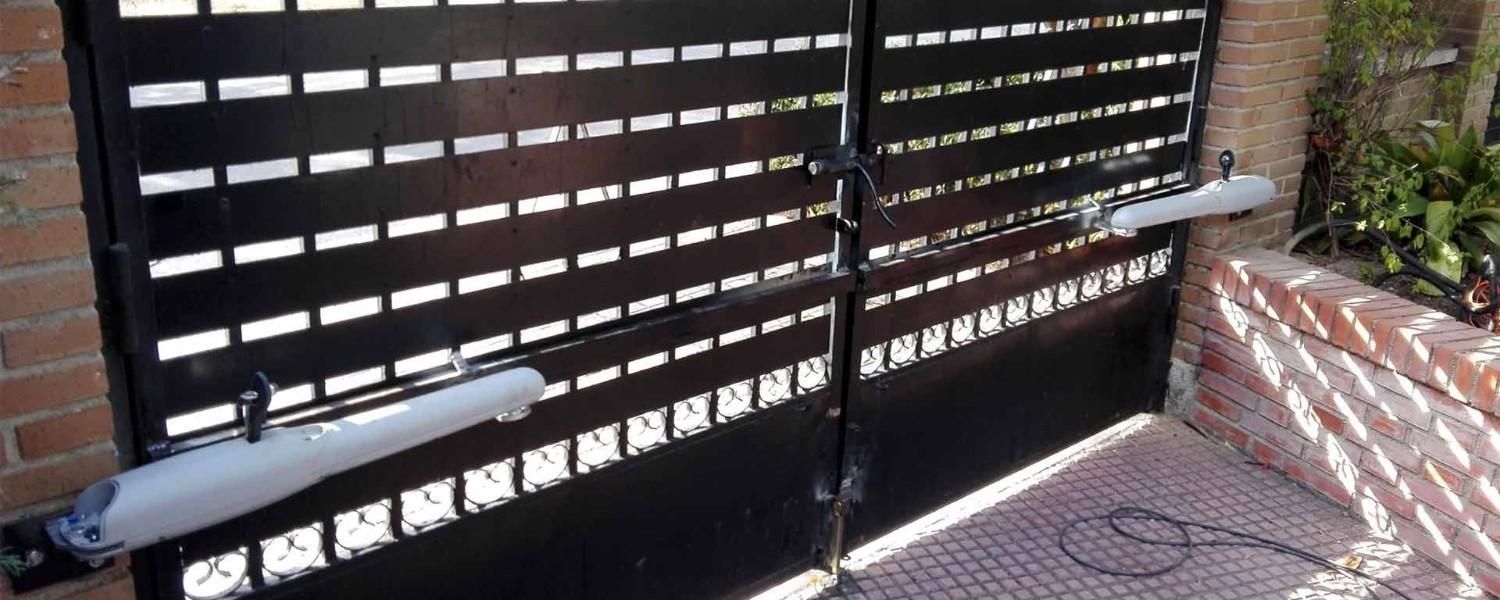 Dos portones de entrada de metal negro con abrepuertas mecánicos automáticos de color claro, fijados a pilares de ladrillo.