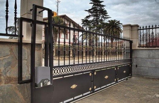 Una puerta corredera de metal negro con un motor gris permanece cerrada frente a un muro de piedra y una casa.