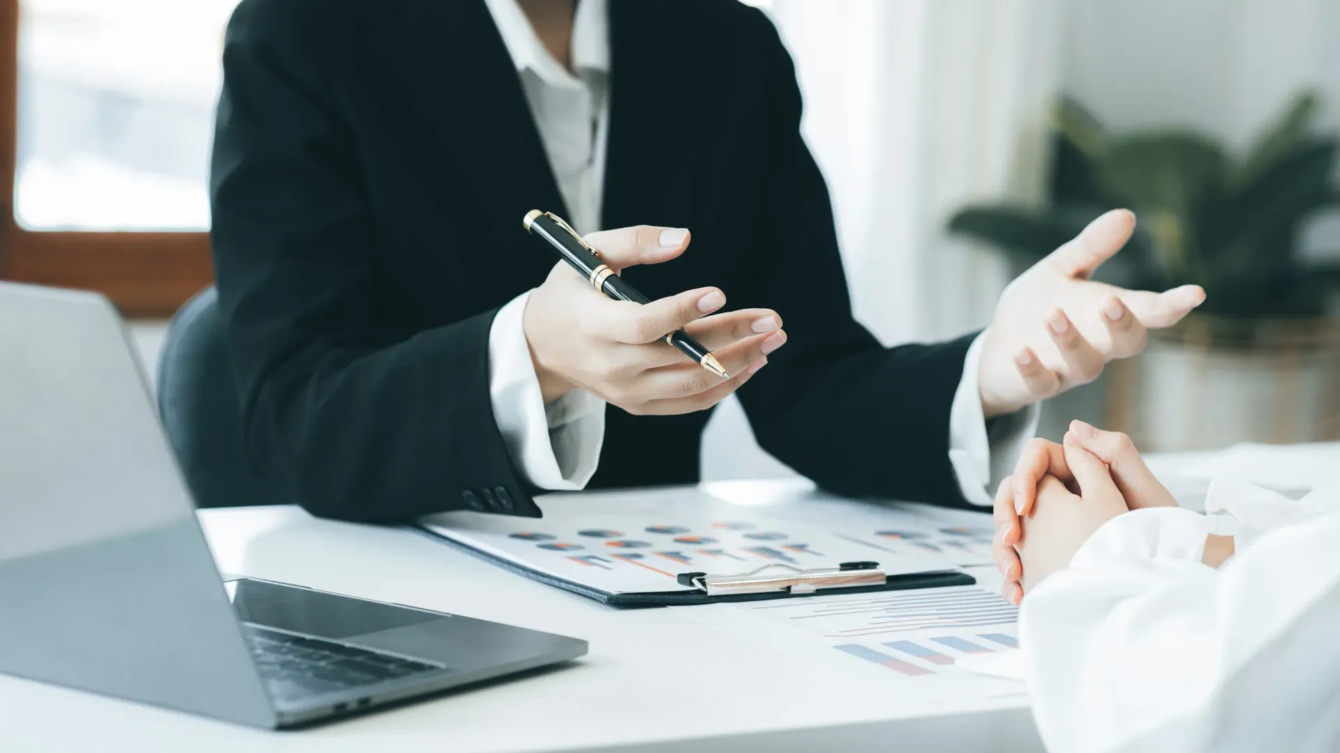 Persona con traje haciendo gestos con un bolígrafo, discutiendo papeles con otra persona en una mesa