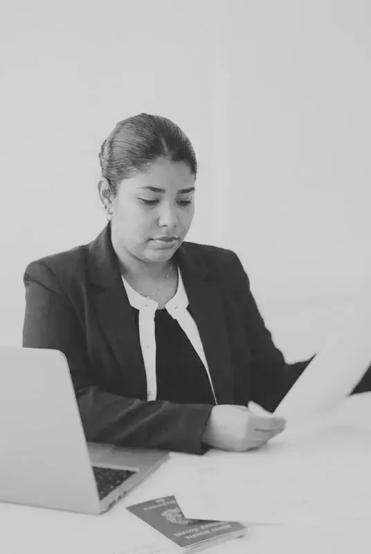 Mujer con blazer revisando documentos en un escritorio con una computadora portátil y un pasaporte.