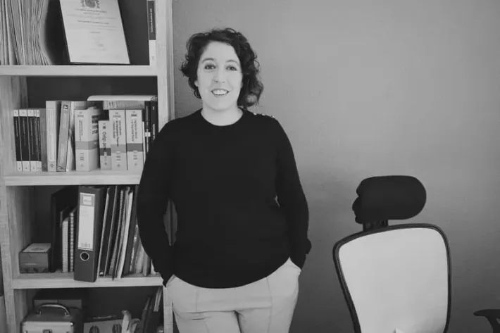 Mujer de pie, sonriendo, con las manos en los bolsillos. Apoyada en una estantería, en un entorno de oficina.