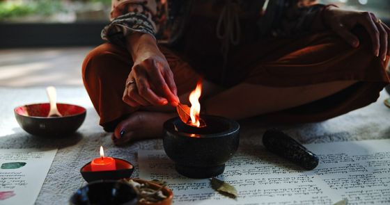 Una mujer con pantalones marrones enciende una hoja sobre una llama en un recipiente negro, rodeada de otras velas y recipientes.