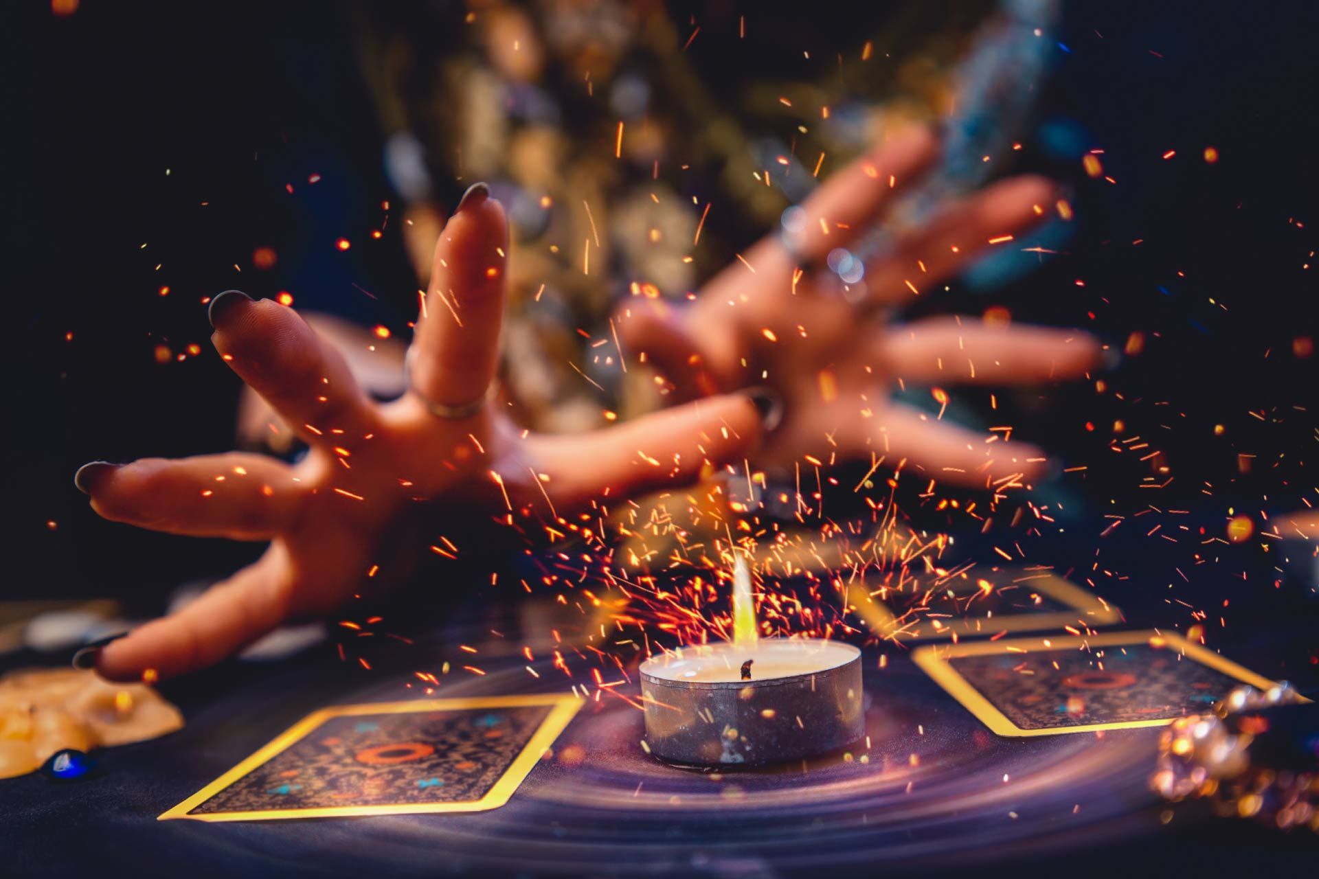 Manos lanzando hechizos sobre cartas del tarot, chispas, vela encendida sobre una mesa oscura.