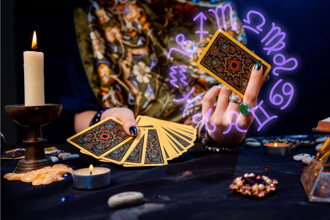 Manos sosteniendo cartas del tarot, rodeadas de velas, símbolos astrológicos y cristales sobre una mesa.
