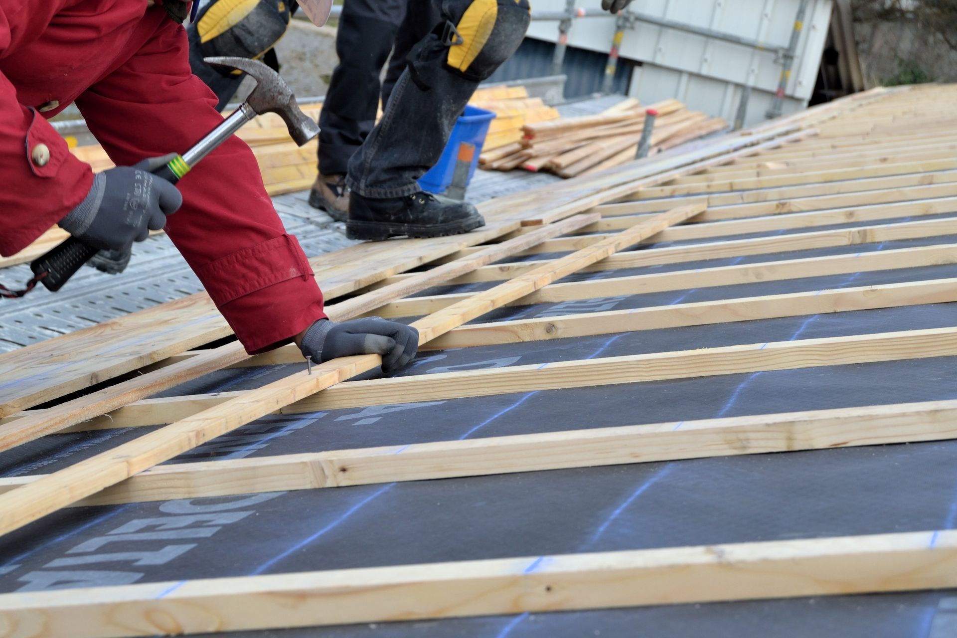 Un ouvrier en veste utilise un marteau pour clouer des lattes de bois sur un toit recouvert d'une sous-couche de protection.