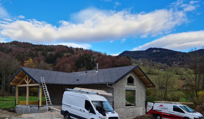 Un chantier de construction avec une maison en construction, deux fourgonnettes de service et une échelle, le tout sur fond de paysage montagneux.