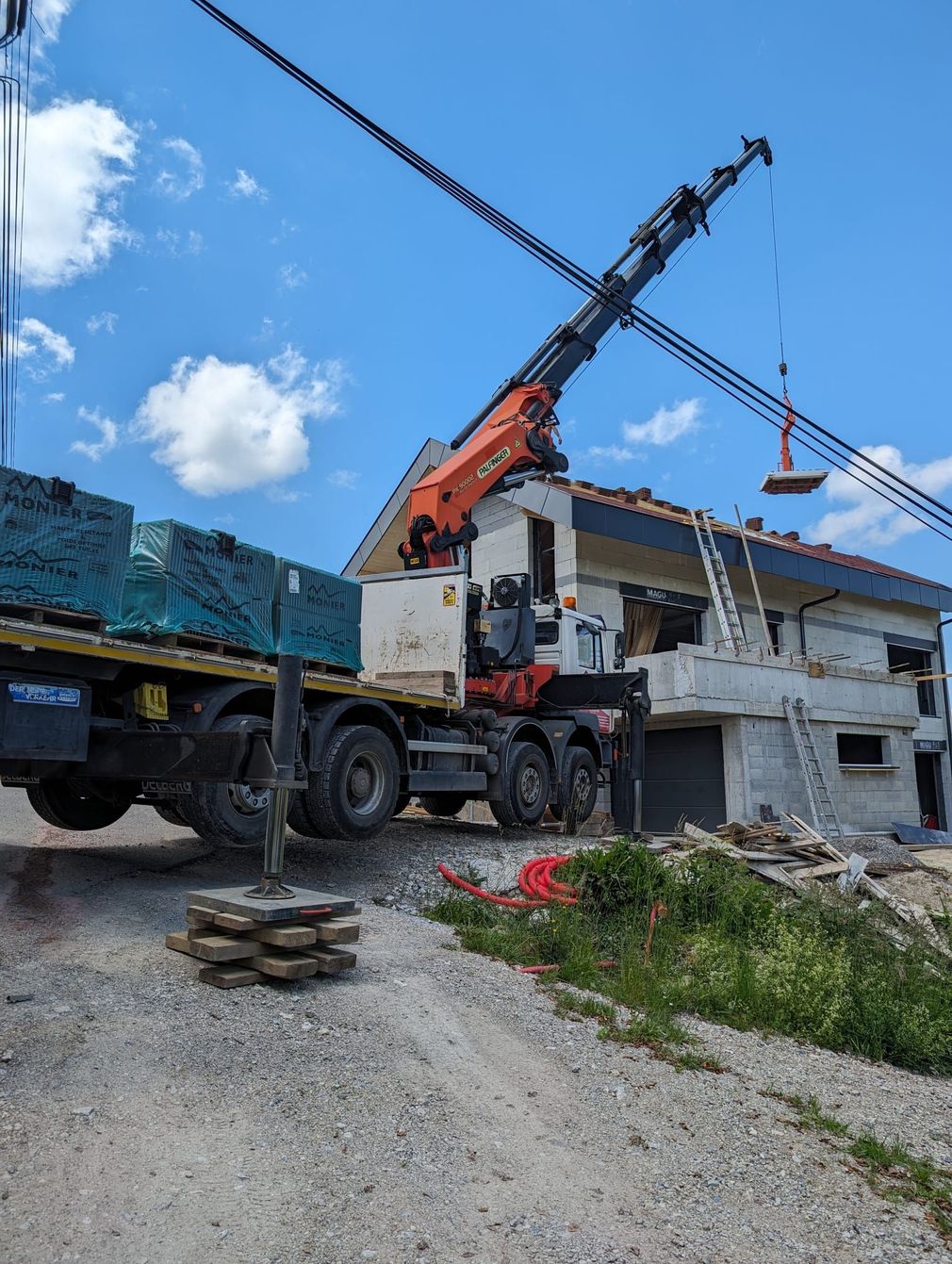 Un camion-grue stationné sur un chantier de construction, en train de soulever une charge pour la déposer sur le toit d'une maison en construction.