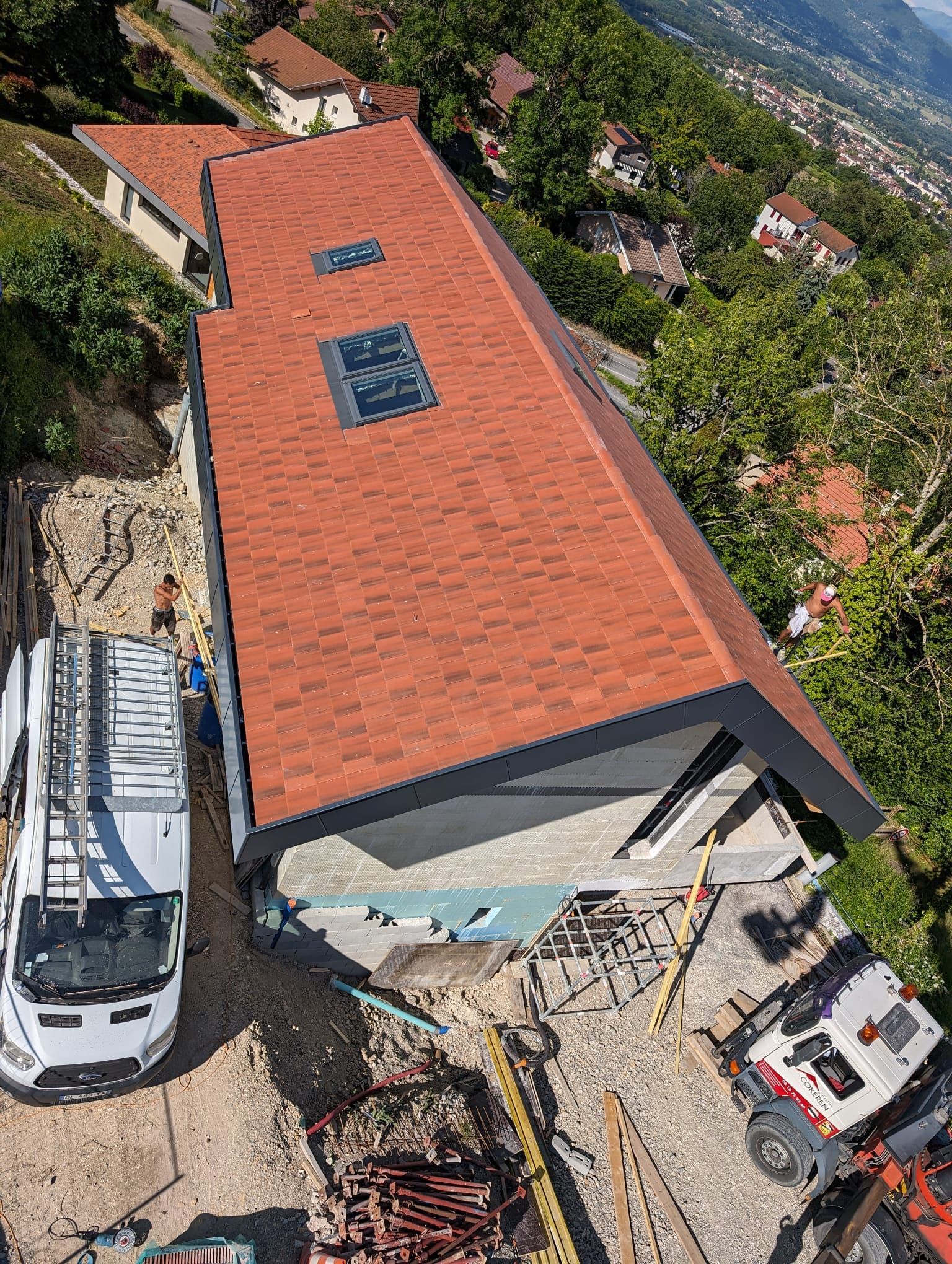 Vue aérienne d'une maison avec un nouveau toit en tuiles, des puits de lumière et des engins de chantier stationnés dans la cour.