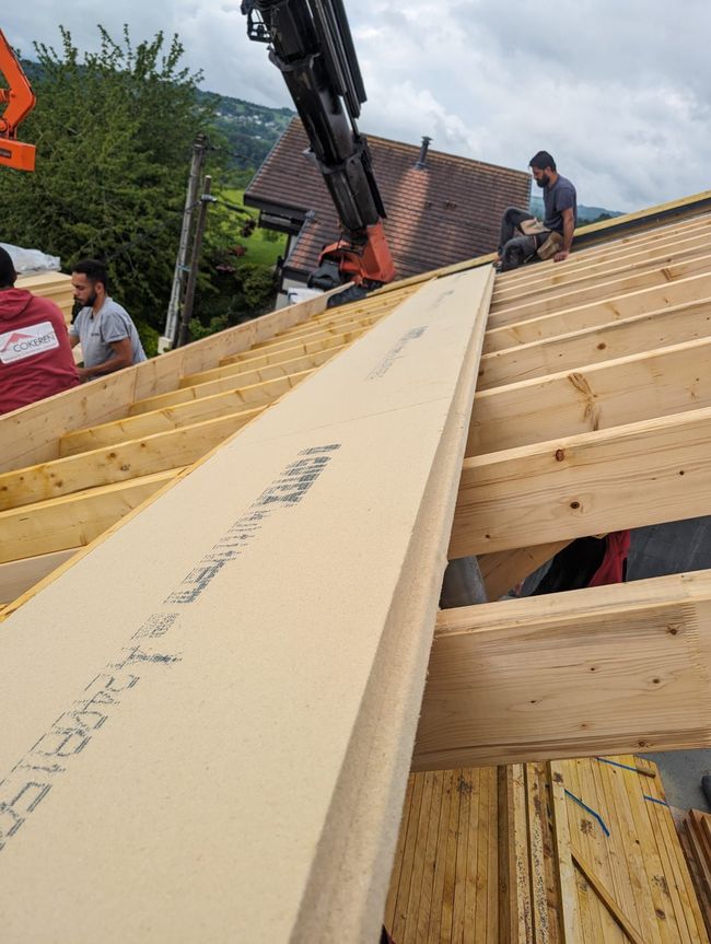 Des ouvriers du bâtiment installent des panneaux de toiture en bois sur la charpente d'un bâtiment, une grue se trouvant à proximité.