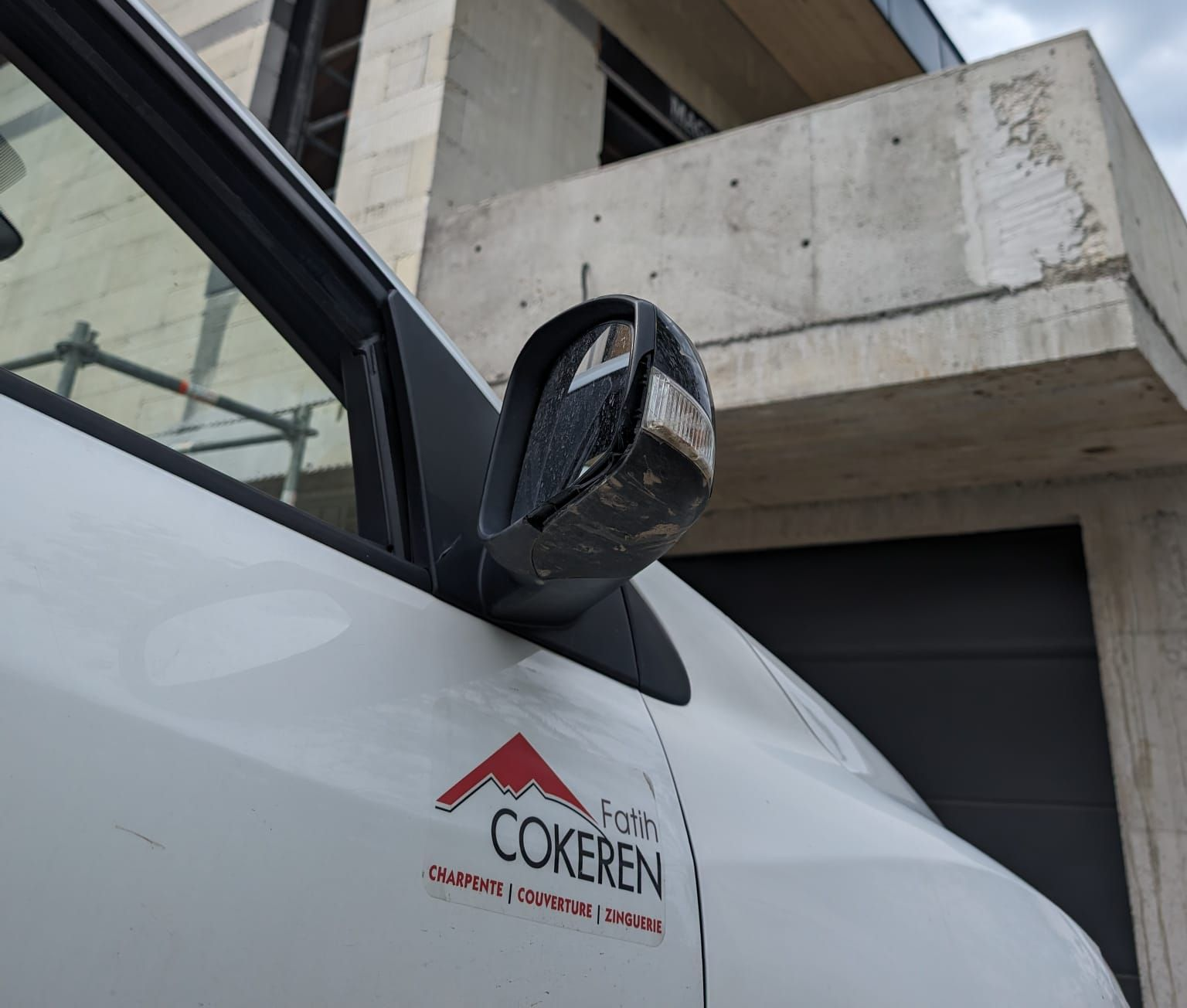 Le rétroviseur latéral d'une camionnette, garée devant un bâtiment moderne en béton.