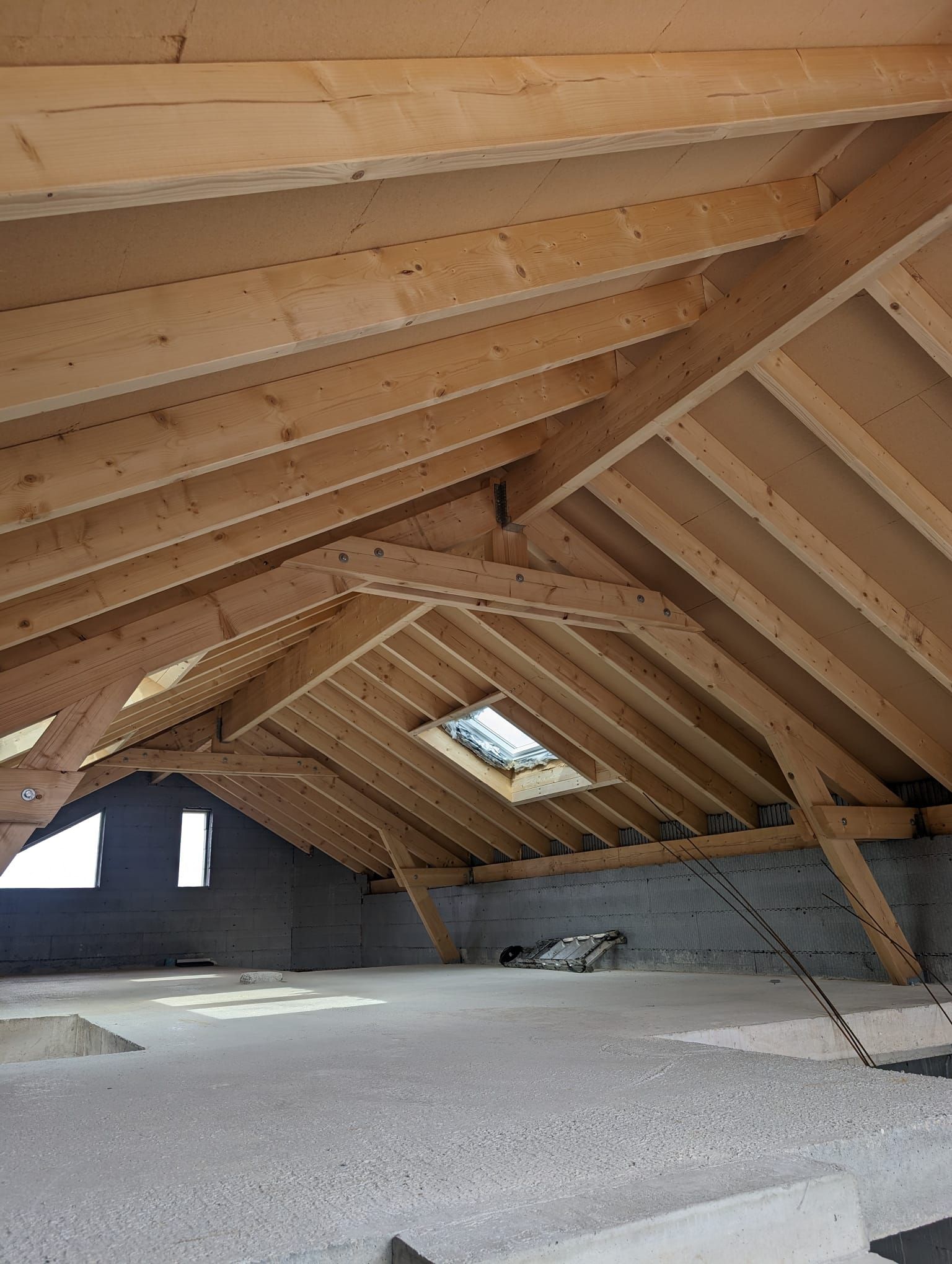 Un intérieur de grenier inachevé avec une charpente apparente, un puits de lumière et un sol en béton.