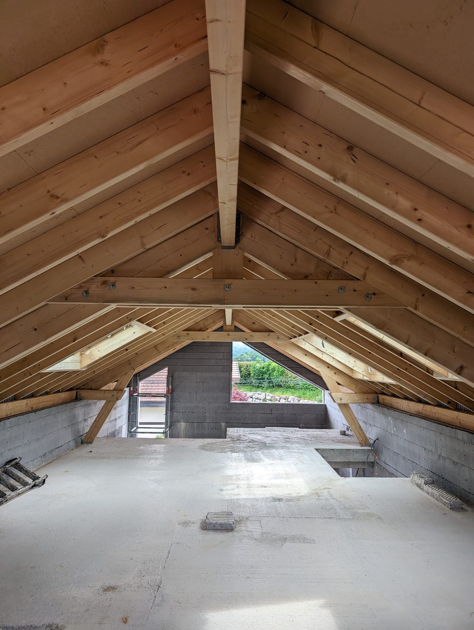 Intérieur de grenier non aménagé présentant des chevrons en bois apparents, une poutre faîtière centrale et un sol en béton avec une fenêtre.