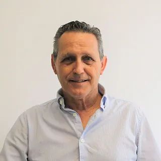 Un hombre con una camisa azul sonríe a la cámara mientras está parado frente a una pared blanca.