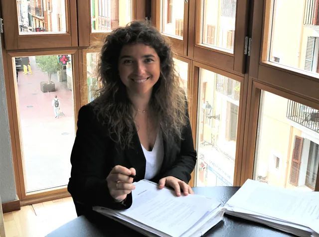Una mujer está sentada en un escritorio frente a una ventana sosteniendo un libro.