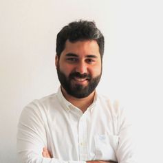 Un hombre con barba lleva una camisa blanca y sonríe con los brazos cruzados.