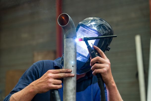 Un homme réalise un soudage à l'arc sur un tube métallique avec une torche de soudure et un masque de sécurité.