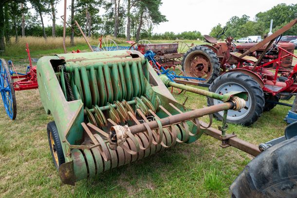 Vieille presse à balles agricole avec dents rotatives, exposée dans un champ aux côtés de tracteurs anciens.