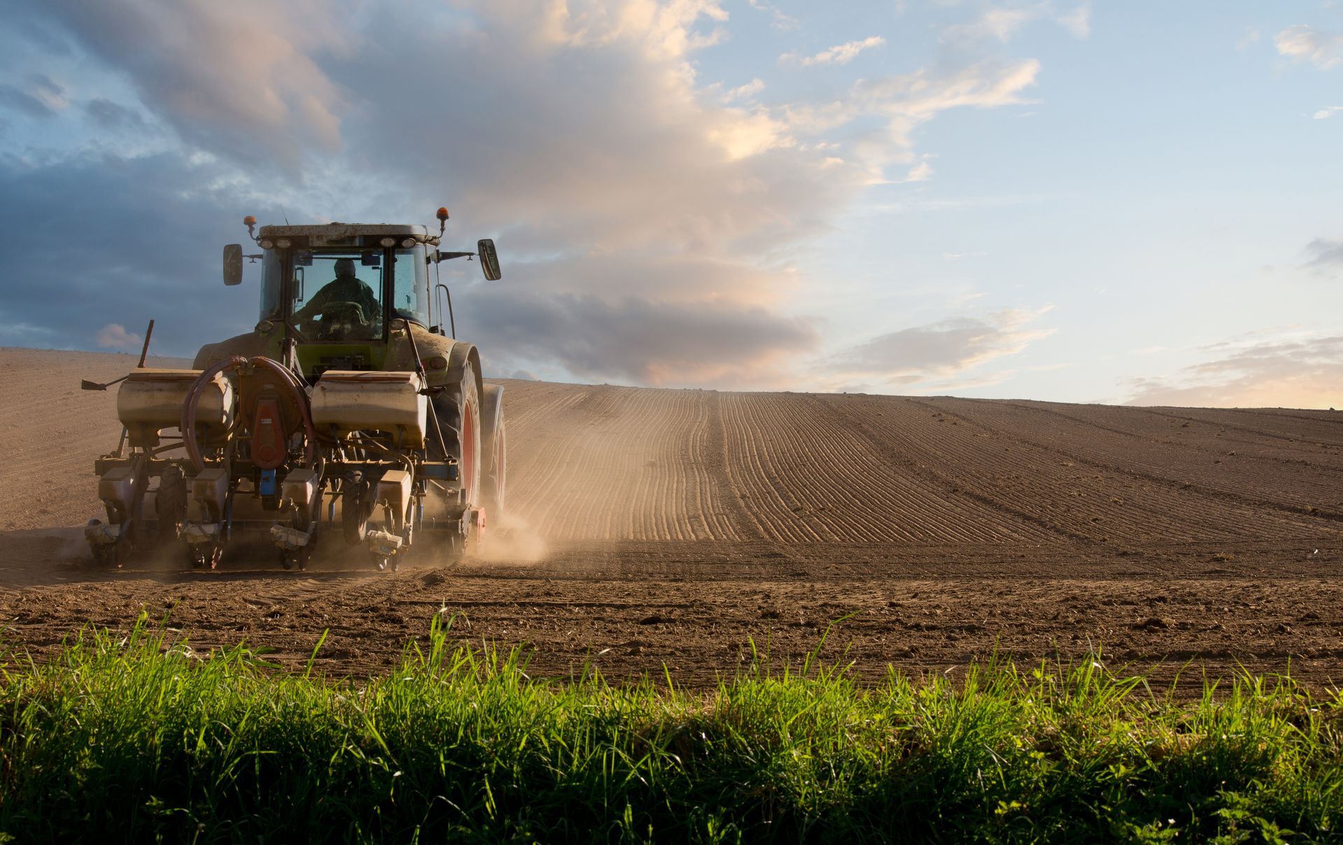 Tracteur agricole en train de semer un champ labouré sous un ciel nuageux au coucher du soleil.