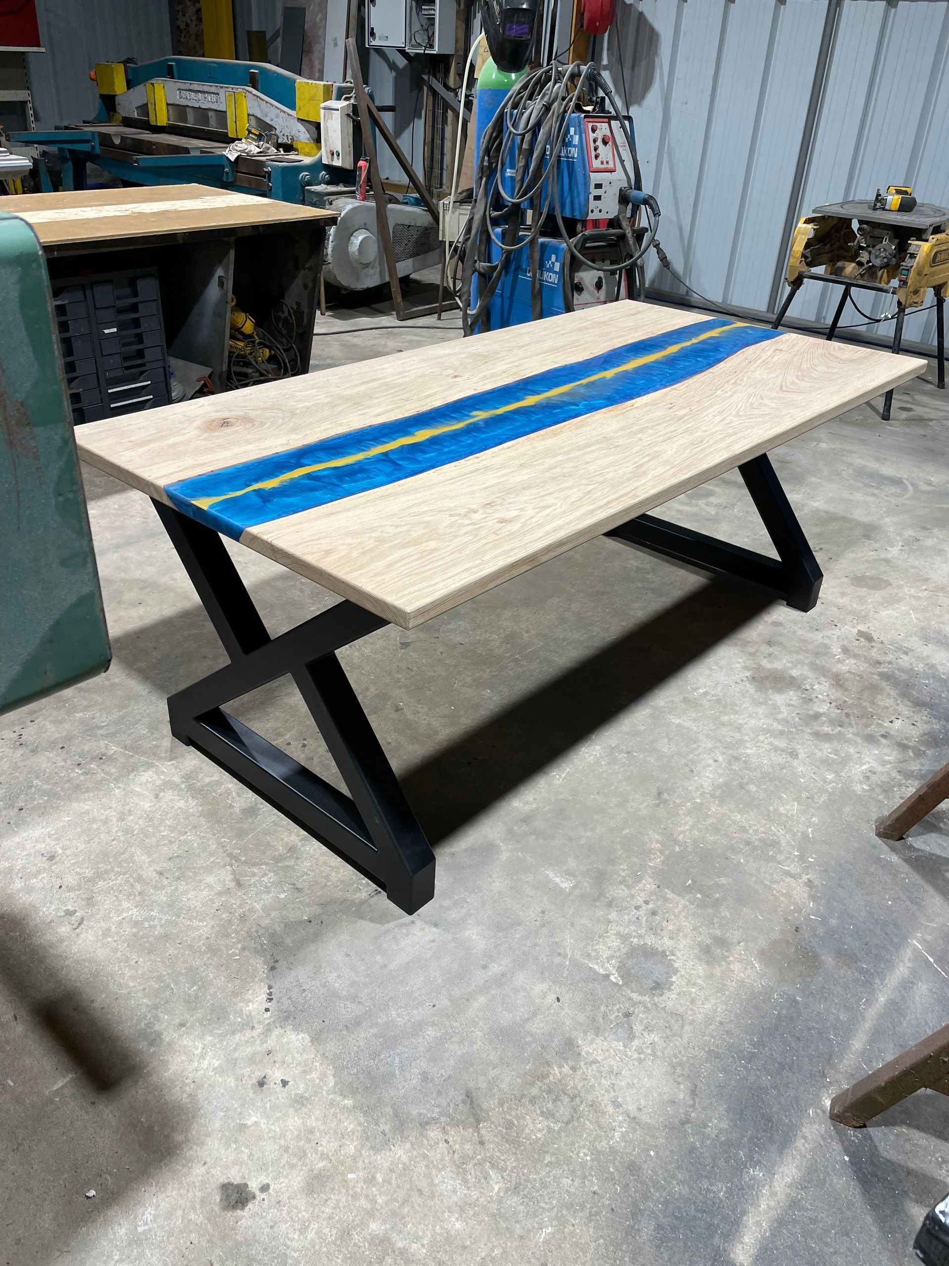 Table en bois avec insert en résine bleue et pieds en métal noir dans un atelier de fabrication.