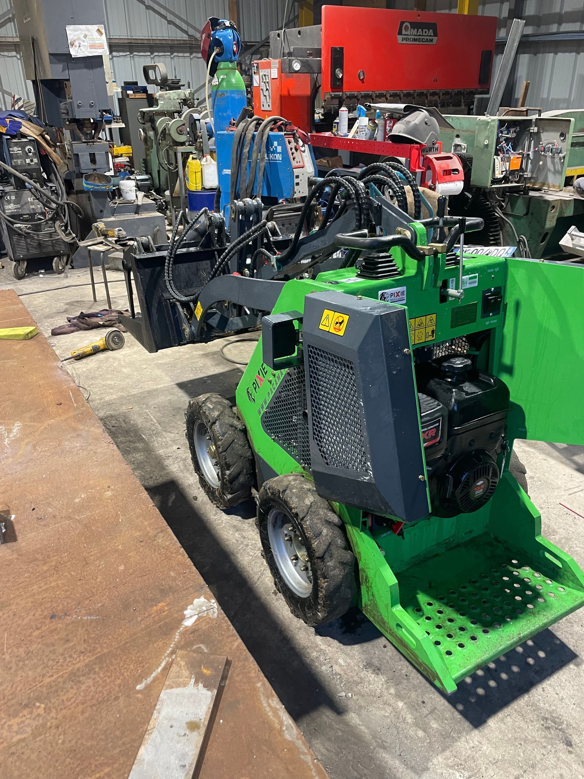 Une chargeuse compacte verte dans un atelier, à côté d'autres machines et outils industriels.