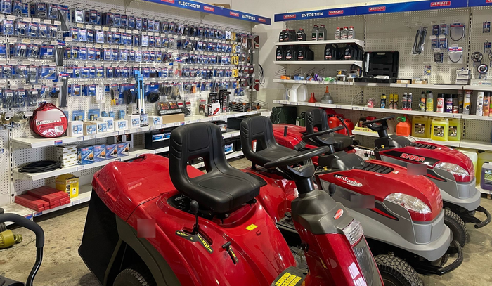 Des tondeuses autoportées rouges et grises sont exposées dans une allée d'un magasin, à côté d'outils de jardinage sur les étagères.