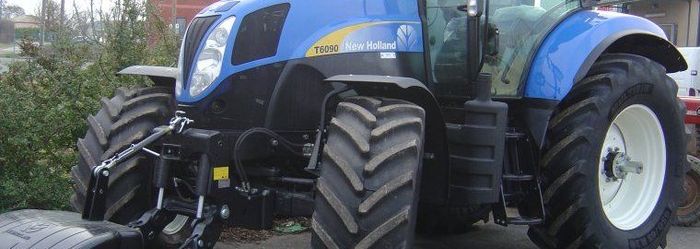Tracteur bleu à gros pneus garé à l'extérieur, près d'un muret en pierre et d'arbres