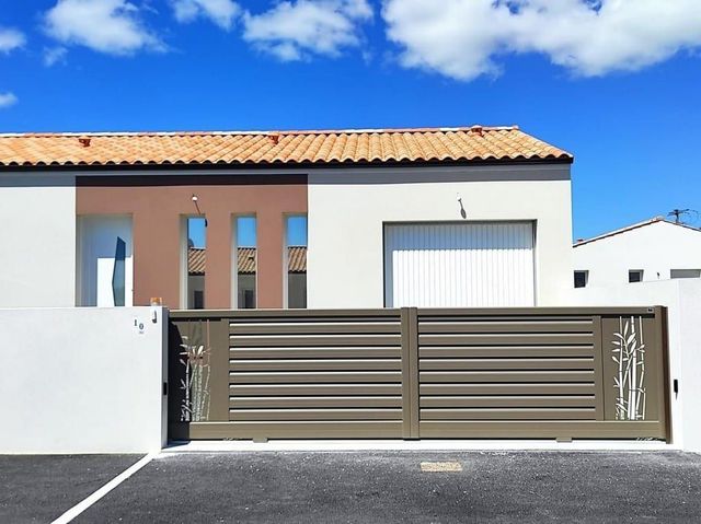 Maison moderne avec toit de tuiles et porte de garage blanche derrière un portail métallique gris sous un ciel bleu