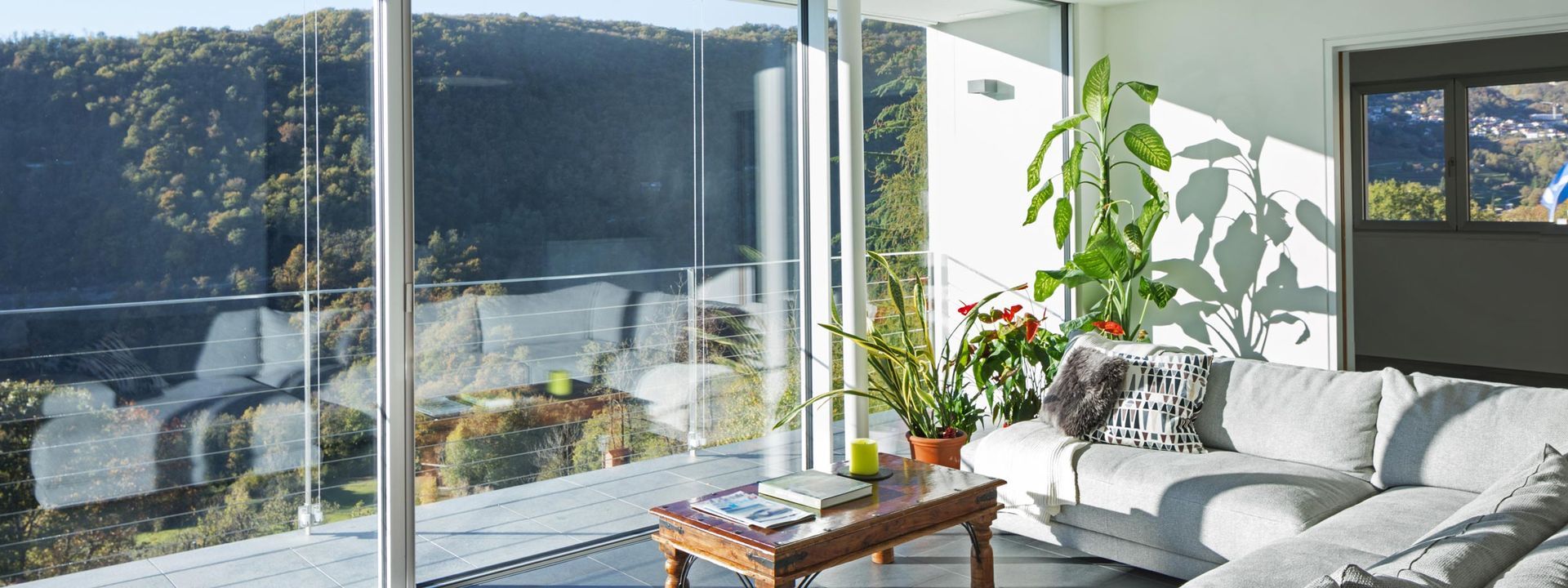 Salon lumineux avec un canapé gris, des plantes en pot et la lumière du soleil qui inonde les pièces à travers de grandes baies vitrées.