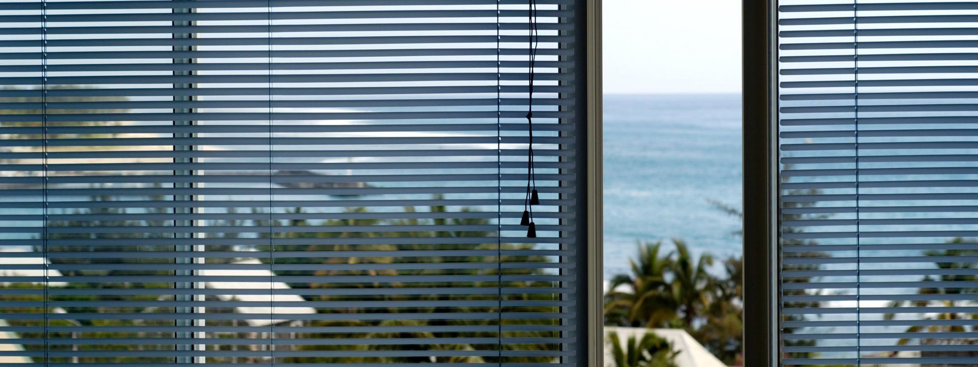 Vue sur l'océan à travers des stores horizontaux, avec des palmiers et des toits au premier plan.