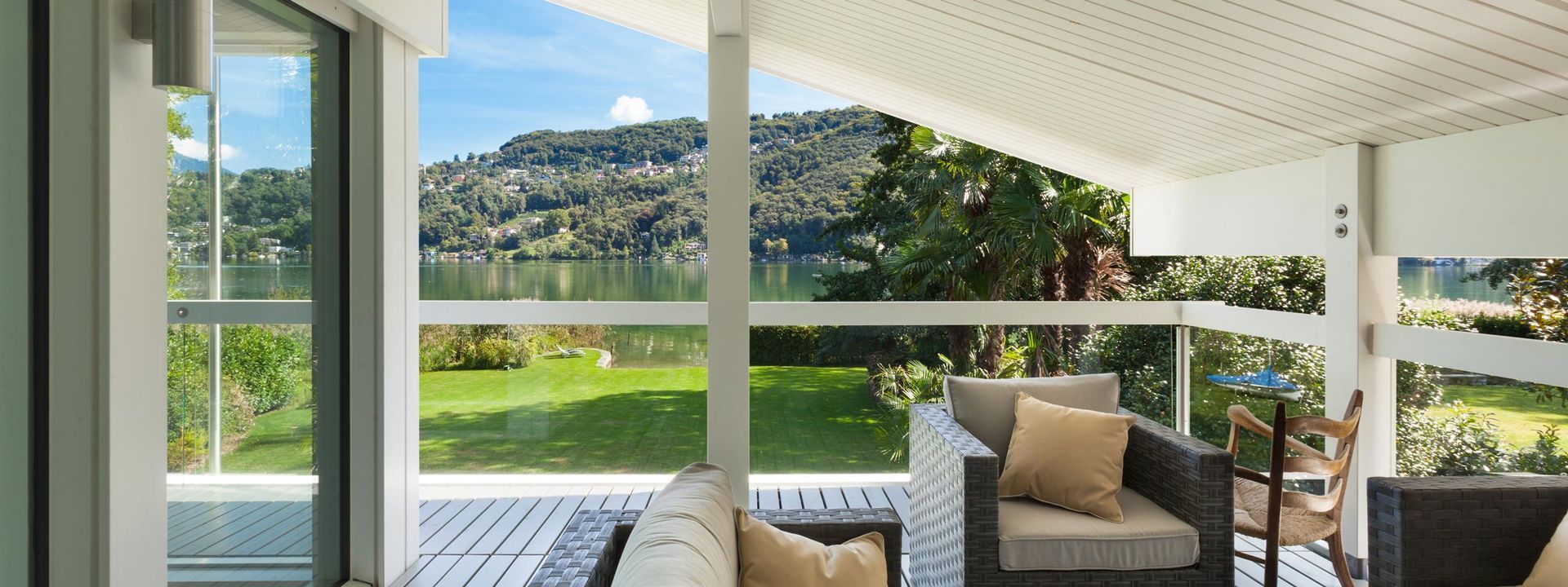 Véranda couverte avec fauteuil en osier, coussins beiges et vue sur une pelouse verdoyante et des arbres