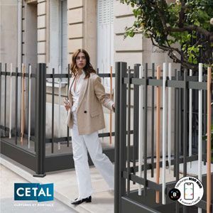 Une femme en blazer beige et pantalon blanc passe devant une clôture métallique à l'extérieur d'un bâtiment.