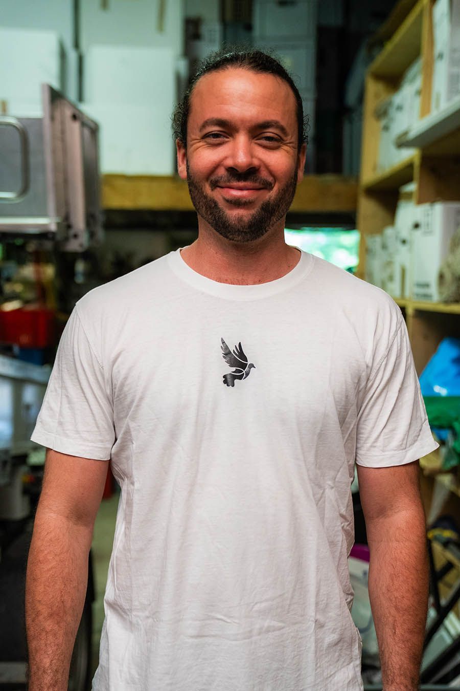 Mann in weißem T-Shirt mit kleinem Emblem, lächelnd im Innenbereich.