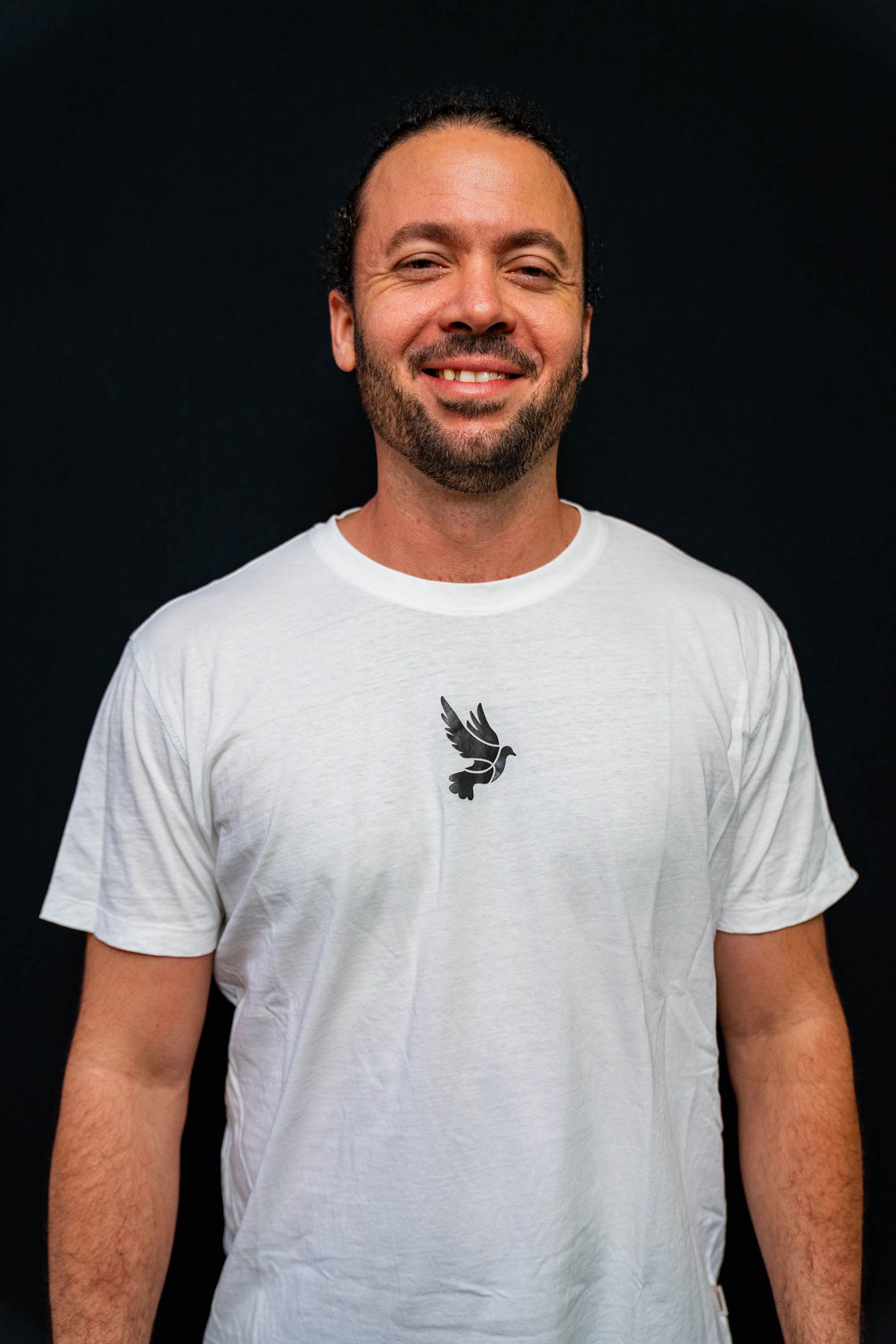 Lächelnder Mann mit Bart, der ein weißes T-Shirt mit einem kleinen Vogelmuster trägt; schwarzer Hintergrund.