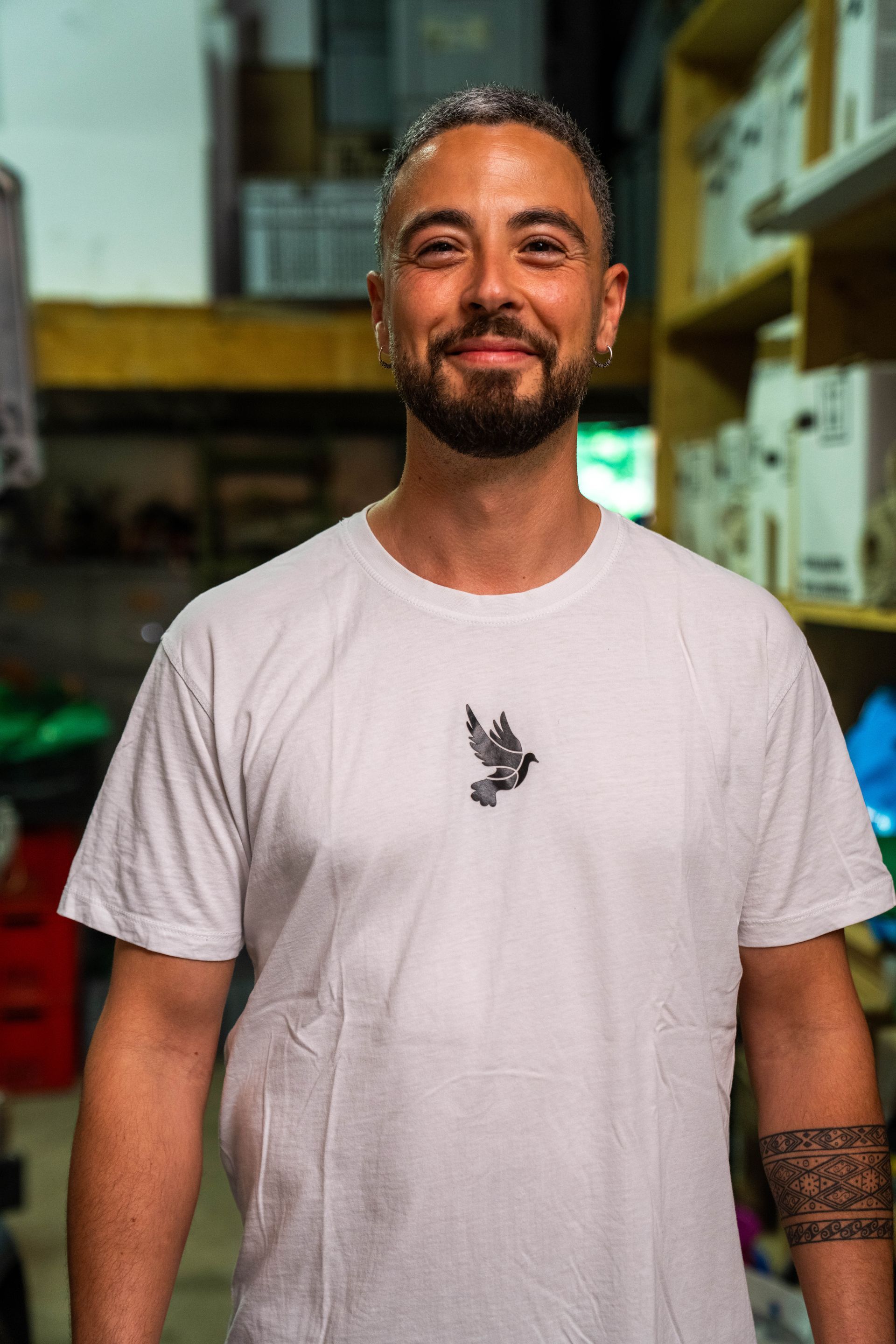 Mann in weißem T-Shirt mit Vogelgrafik lächelt in einem Lagerhaus.