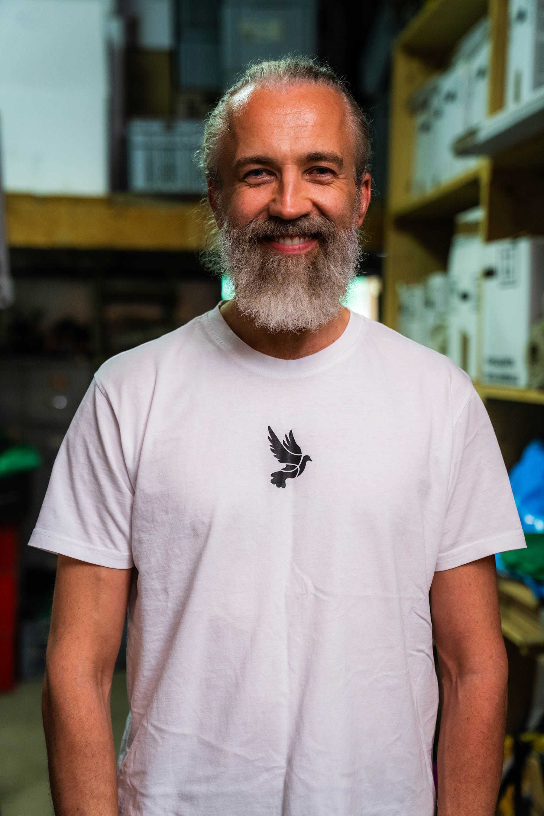 Mann mit langem grauem Bart lächelt und trägt ein weißes T-Shirt. Steht in einem überfüllten Raum.