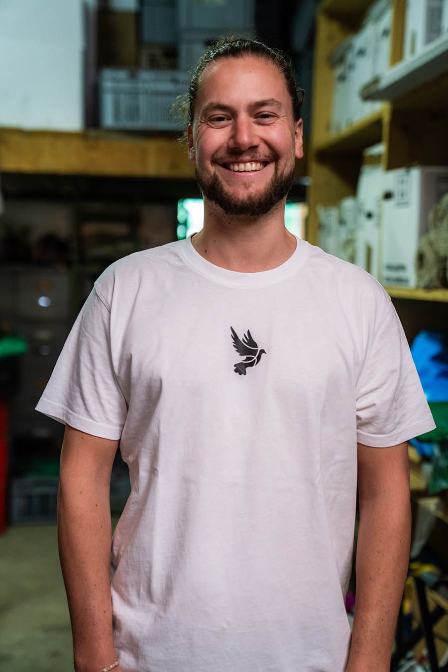 Lächelnder Mann mit Bart und weißem T-Shirt mit Vogelmotiv steht in einem Lagerraum.