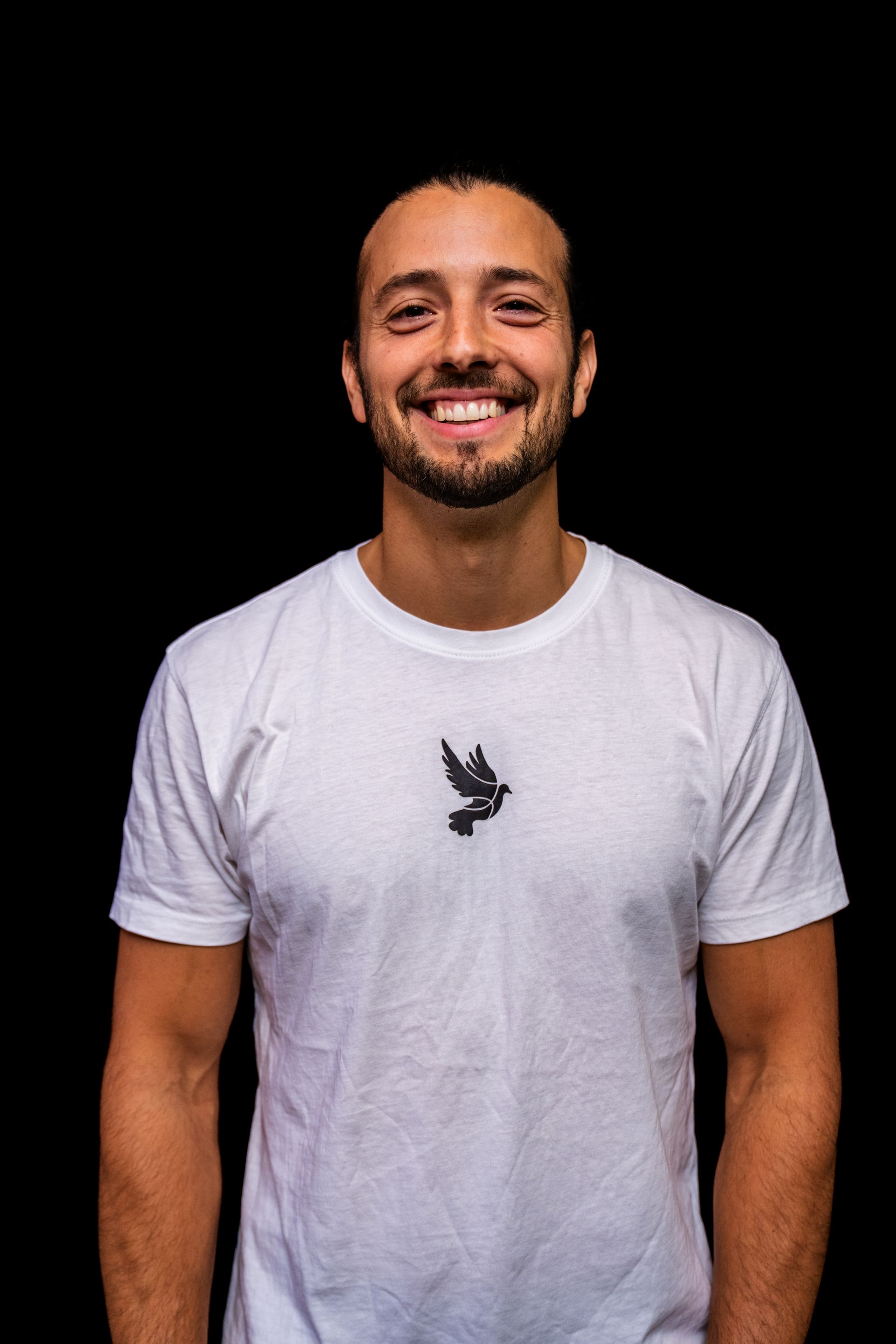 Lächelnder Mann in weißem T-Shirt mit Vogellogo vor schwarzem Hintergrund.