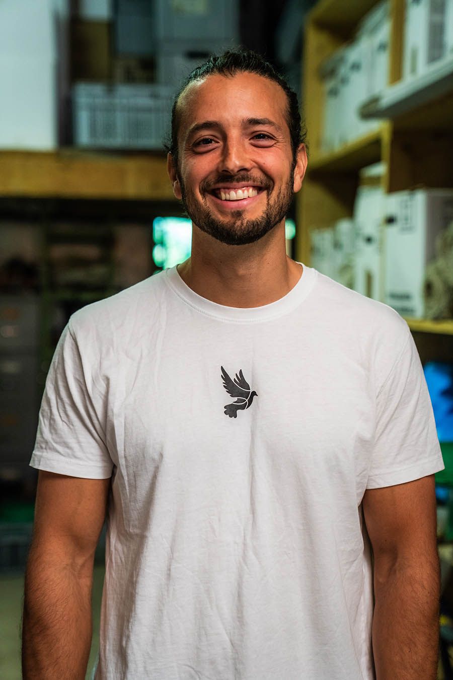 Lächelnder Mann mit Bart, der ein weißes T-Shirt mit Vogelaufdruck trägt und in einer Lagerhalle steht.