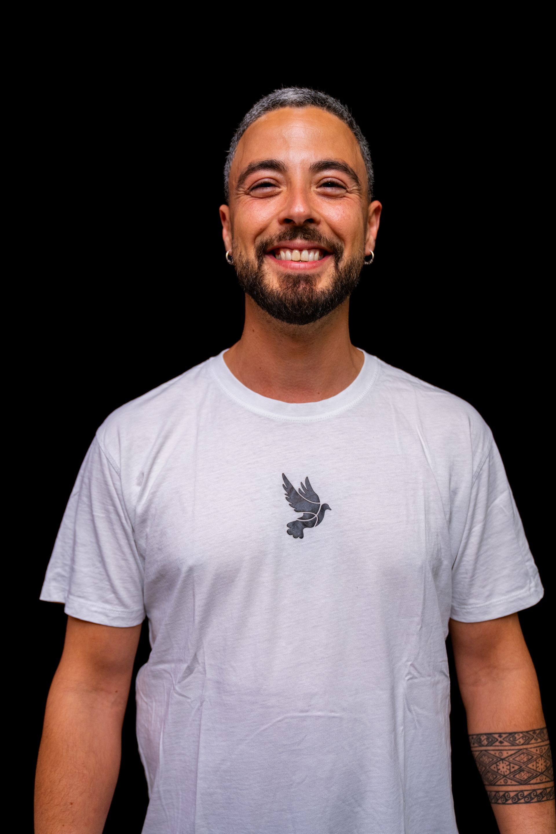 Mann mit kurzem Bart, lächelnd. Trägt ein weißes T-Shirt mit einem kleinen Taubenmuster auf schwarzem Hintergrund.